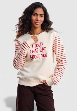 Bluza z kremowym tułowiem, czerwonym napisem „I TOLD CHAT GPT ABOUT YOU” oraz czerwono-białymi paskowanymi rękawami. Bawełniany materiał, luźny krój.