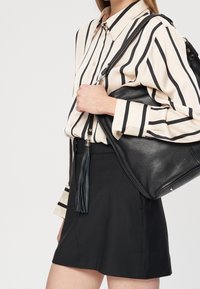 Donna che indossa una gonna nera e una camicia beige con righe nere, porta una borsa a tracolla in pelle nera con dettaglio a frange.