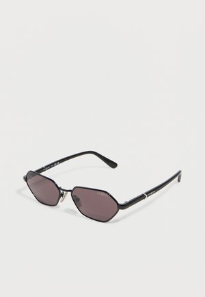 Gafas de sol hexagonales con montura de metal negra, lentes de color oscuro y marca "Vogue" en las patillas y lentes.