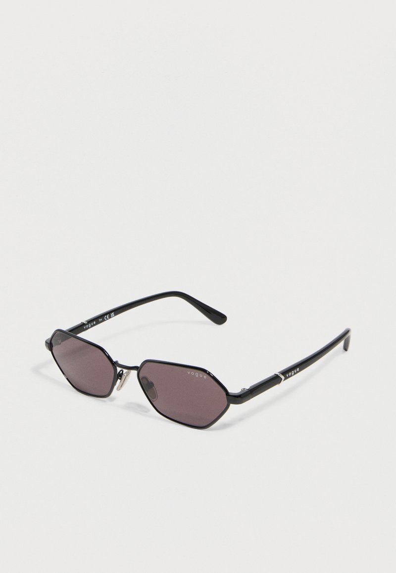 Μαύρα εξαγωνικά γυαλιά ηλίου με μεταλλικό σκελετό, σκούρους φακούς και το λογότυπο "Vogue" στα πλαϊνά και τους φακούς.