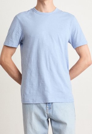 Homme portant un t-shirt uni bleu clair à manches courtes et col rond, et un jean bleu clair, debout les mains derrière le dos sur un fond neutre.