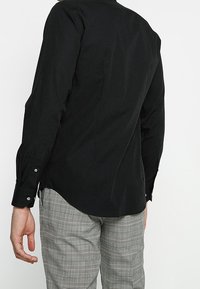 Black long-sleeve shirt with a smooth texture, flat back seam details, and button cuffs. Paired with grey plaid trousers.