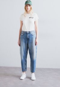 Λευκό t-shirt με κείμενο "POLO", ανοιχτό μπλε ψηλόμεσο τζιν, πράσινο καπέλο και λευκά sneakers, με χαλαρή εφαρμογή και ελαφριά ξεθωριασμένη όψη.