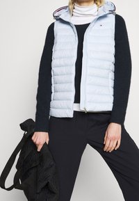 Personne portant un gilet matelassé bleu clair sur un pull noir et un col roulé blanc, tenant un grand sac matelassé noir d'une main.