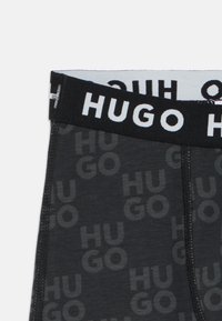 Juodos bokso kelnaitės su tamsiai pilku pakartojančiu „HUGO“ logotipu. Yra juodas elastingas juosmuo su baltu užrašu ir kontrastingomis siūlėmis.