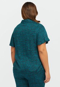 Top a maniche corte in teal con motivo a pois neri sparsi. Tessuto liscio, vestibilità rilassata e dettaglio con chiusura a bottoni sul retro del collo.