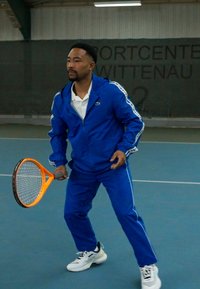 Kék melegítő szett fehér csíkokkal, fehér pólóing és sportcipők; egy narancssárga teniszütőt tart a kezében egy fedett pályán.