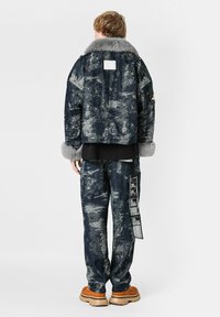 Chaqueta navy con cuello de piel sintética, diseño texturizado y acentos grises; combinada con pantalones a juego que presentan gráficos y una etiqueta colgante.
