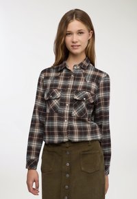 Giovane donna con indosso una camicia marrone a quadri abbottonata, infilata in una gonna verde oliva con bottoni frontali e tasche anteriori.