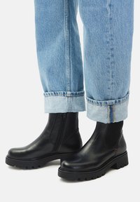 Čierne kožené členkové topánky s hrubou podrážkou, kombinované s svetlomodrými riflami s manžetami na bielom pozadí.