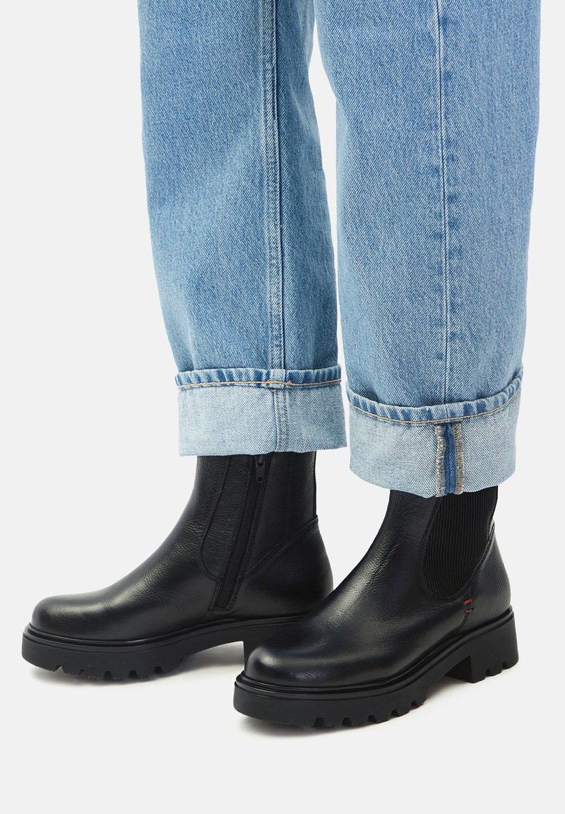 Čierne kožené členkové topánky s hrubou podrážkou, kombinované s svetlomodrými riflami s manžetami na bielom pozadí.