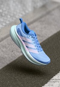 Chaussure de sport bleu clair avec dessus en maille, trois bandes roses sur le côté, et une semelle épaisse vert menthe. Présente un détail texturé.