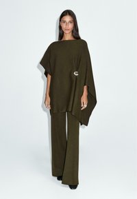 Poncho en laine vert olive avec un ourlet asymétrique, orné d'une broche argentée décorative et accompagné d'un pantalon à jambes larges assorti.
