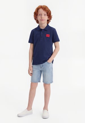 Garçon roux debout, une main dans la poche, portant un polo bleu marine, un short en jean clair et des chaussures blanches slip-on.