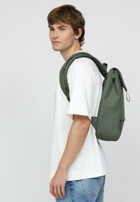 Groene rugzak gemaakt van duurzaam materiaal met een oprolbare sluiting en verstelbare schouderbanden, gedragen door een persoon in een wit shirt en jeans.