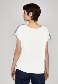 Camiseta blanca de manga corta con cuello redondo y detalles con estampado floral en las mangas. Tela suave, ajuste relajado y corte a la altura de la cadera.