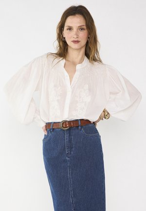 Donna che indossa una camicetta bianca ricamata con maniche a sbuffo infilata in una gonna di jeans a righe blu, accessoriata con una cintura marrone.