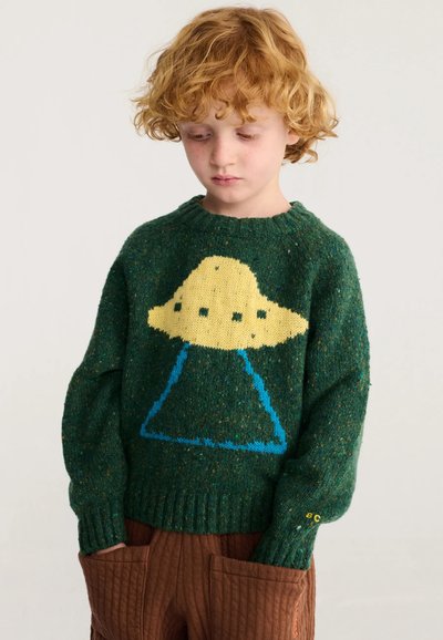 Enfant aux cheveux roux bouclés portant un pull vert avec un motif OVNI jaune et un rayon bleu, associé à un pantalon matelassé marron avec de grandes poches.