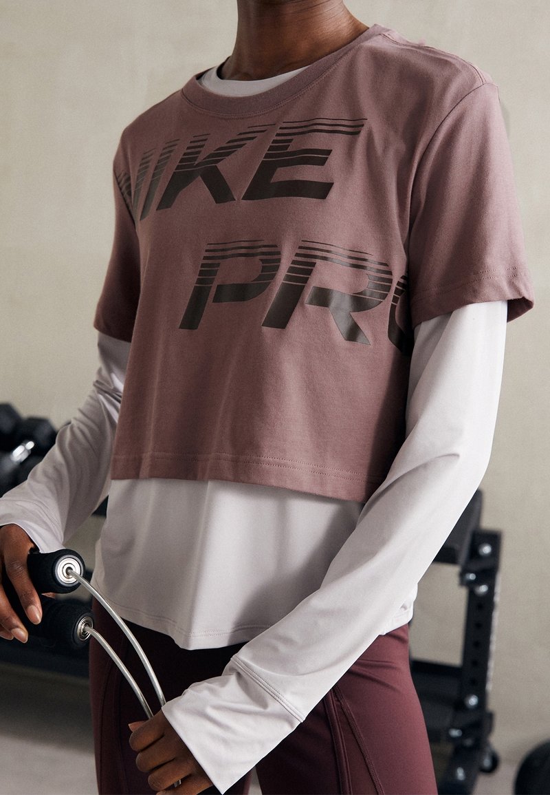 Lyhyet hihat, lyhennetty, mauve urheilupaita, jossa on tummaa harmaata väriä käytetty tekstissä "NIKE PRO", pitkähihaisen valkoisen paidan päällä, pidetty käsissä liikuntavälineiden kanssa.