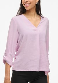 Blusa viola chiaro con scollo a V, maniche a tre quarti arrotolate, tessuto texture e vestibilità ampia. Indossata con pantaloni neri.