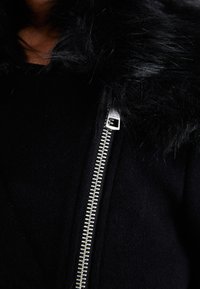 Čierna bunda s umelým kožušinovým golierom, obsahuje strieborný zips a mäkkú textúrovanú tkaninu. Detailný záber zvýrazňuje zips a textúru goliera.