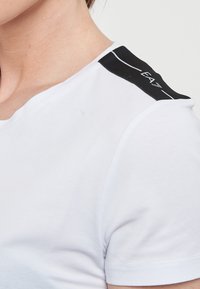 T-shirt bianco a maniche corte con un accento nero sulla spalla che presenta un logo. Tessuto morbido e liscio con un design aderente.