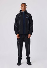 Cazadora negra con detalles en azul, con capucha y cierre de cremallera, combinada con pantalones negros a juego y calzado negro texturizado.