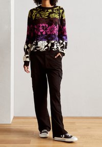 Personne portant un pull multicolore à motif floral, un pantalon cargo noir et des baskets noires, debout sur un sol en bois devant un mur blanc.