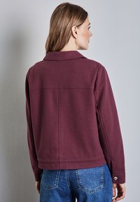 Burgunderfarbene Jacke mit Kragen, langen Ärmeln und lockerer Passform. Verfügt über Nähdetails und Knopfakzente an den Ärmelbündchen. Getragen mit blauen Jeans.