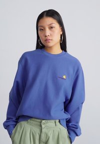 Fialový crewneck sweatshirt s uvolněným střihem, se malým žlutým a vínovým logem na hrudi. V kombinaci se zelenými plisovanými kalhotami.