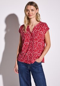 Blusa rossa a maniche corte con scollatura a V, decorata da un motivo astratto bianco. Realizzata in tessuto leggero, ha una vestibilità rilassata e leggere arricciature sulle spalle.