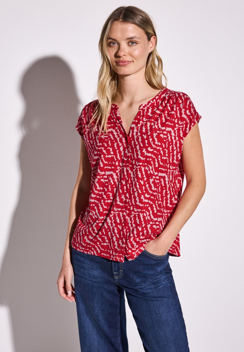 Blusa rossa a maniche corte con scollatura a V, decorata da un motivo astratto bianco. Realizzata in tessuto leggero, ha una vestibilità rilassata e leggere arricciature sulle spalle.