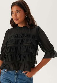 Blusa nera in chiffon con volant sovrapposti, scollo rotondo e maniche a tre quarti a palloncino. Abbinata a jeans in denim azzurro chiaro.