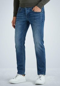 Jeans de mezclilla azul oscuro con un ajuste recto, que cuentan con cinco bolsillos, un ligero desgaste y costuras visibles. Combinados con zapatillas blancas.