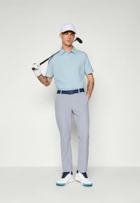 Helehall sinine golfipolo väikese mustriga, hallid püksid ja valged tossud, millel on sinised aktsendid. Modell hoiab golfikeppi.