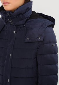 Zbliżenie na osobę w granatowej pikowanej kurtce puchowej z wysokim kołnierzem, klamrą, napami i odczepianym kapturem.