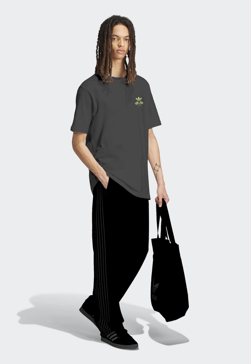 Jongere met dreadlocks draagt een zwarte Adidas trainingsbroek, een donkergrijs T-shirt met logo, zwarte sneakers en houdt een zwarte Adidas schoudertas vast.