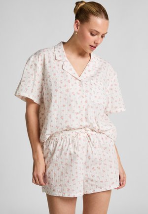 Vrouw in een lichtroze bloemen short-sleeve knoopshirt en bijpassende shorts, staande tegen een effen achtergrond, kijkt naar beneden.