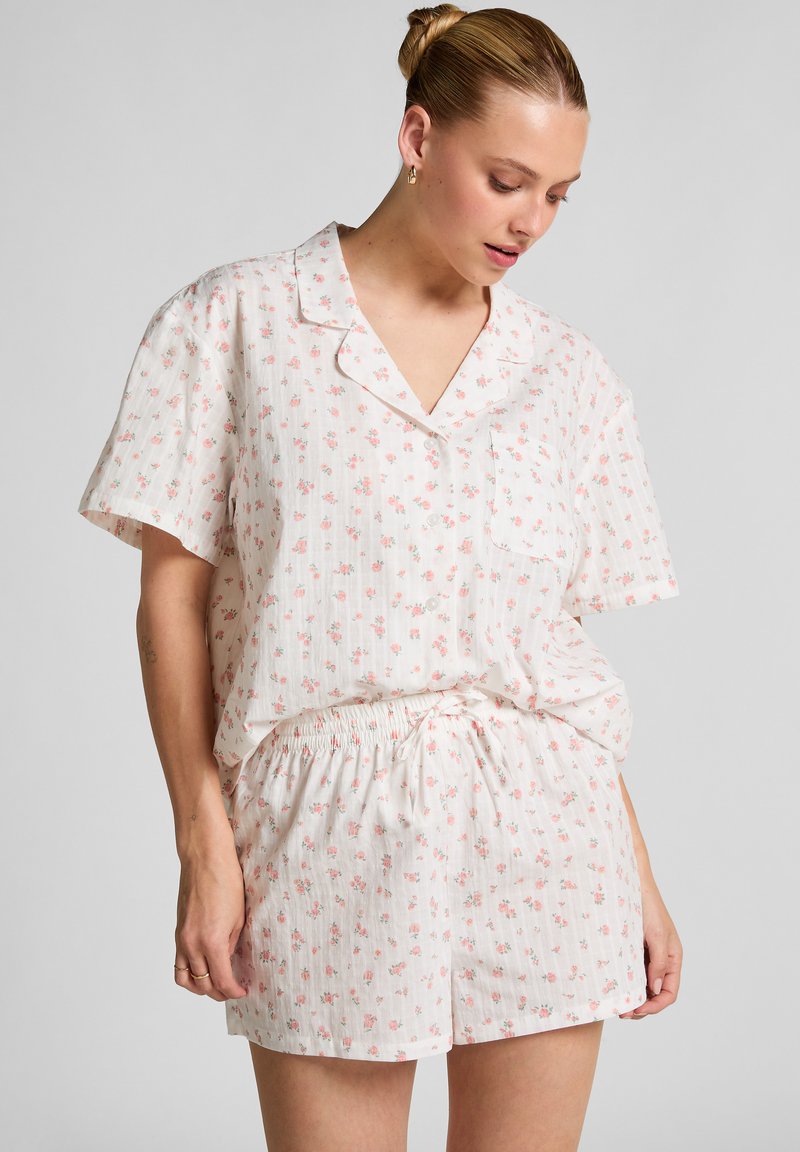 Vrouw in een lichtroze bloemen short-sleeve knoopshirt en bijpassende shorts, staande tegen een effen achtergrond, kijkt naar beneden.