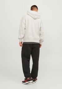 Sweat à capuche gris clair avec une grande capuche, manches côtelées, porté sur un t-shirt noir, assorti à un pantalon de survêtement noir et des baskets avec des accents rouges.