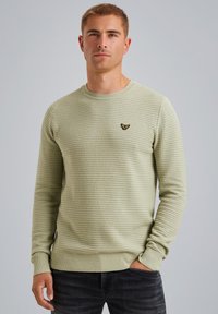 Suéter de punto verde claro con un patrón texturizado, cuello redondo y puños acanalados. Presenta un pequeño logo negro en el pecho.