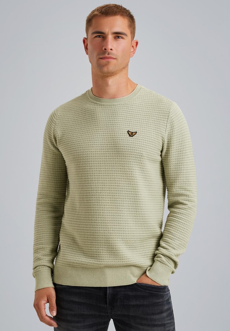 Suéter de punto verde claro con un patrón texturizado, cuello redondo y puños acanalados. Presenta un pequeño logo negro en el pecho.