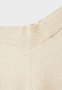Gros plan sur le col d'un pull en maille côtelée beige montrant la texture et les détails des coutures.