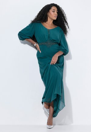 Teal lange jurk met ballonmouwen, voorzien van een met kralen versierde taille. Gecombineerd met zilveren hoge hakken, op een witte achtergrond.