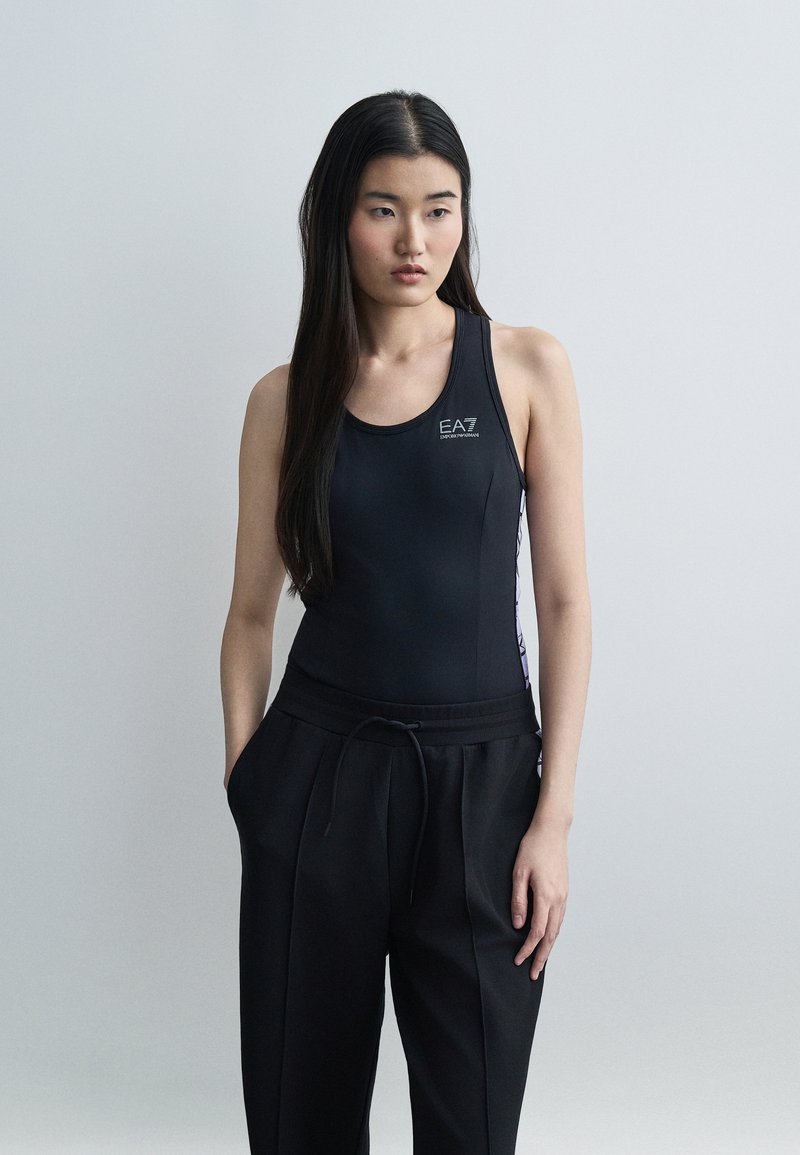 Γυναίκα με μακριά μαύρα μαλλιά, φορώντας μαύρο αμάνικο αθλητικό τοπ EA7 και μαύρο παντελόνι με κορδόνι, όρθια μπροστά σε ένα απλό ανοιχτό γκρι φόντο.