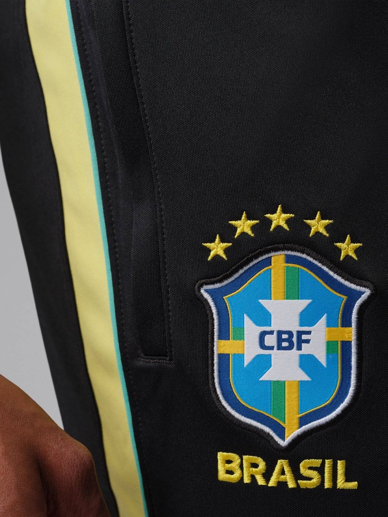 Close-up van zwarte sportkleding met het CBF-embleem van Brazilië, vijf gouden sterren erboven en een gele zijstreep met turkooiskleurige rand.