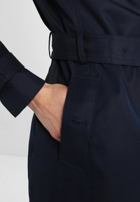 Marinblå trenchcoat med skärp, utrustad med sidficka, slät textur och knäppta manschetter. Tyget är glänsande med skräddarsydda designdetaljer.