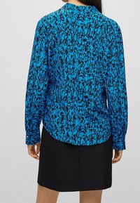 Kvinna iklädd en långärmad blåmönstrad blus och en svart kjol, sedd bakifrån mot en enkel grå bakgrund.