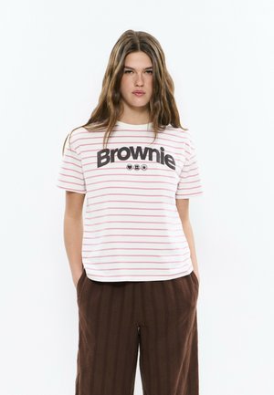 Jeune femme aux cheveux longs portant un T-shirt blanc à rayures roses avec le texte "Brownie" et un pantalon large marron, les mains dans les poches.