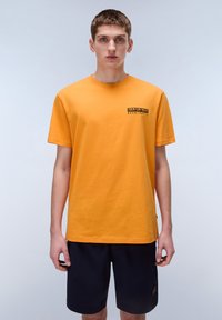Oranssi puuvillainen t-paita lyhyillä hihoilla, pyöreä kaula-aukko ja pieni musta painettu logo vasemmassa rinnassa. Yhdistetty tummiin shortseihin.
